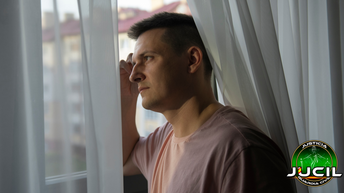 Hombre de perfil, con expresión pensativa, mirando por la ventana entre cortinas blancas. Lleva una camiseta clara y aparece el logotipo de JUCIL en la parte inferior derecha, símbolo de la asociación profesional de la Guardia Civil.
