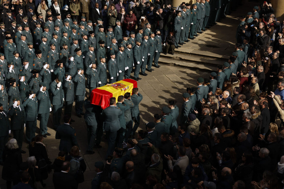 Honor fúnebre de la Guardia Civil con un féretro cubierto por la bandera de España, portado por agentes formados en fila y rodeado de compañeros y ciudadanos que rinden homenaje solemne.