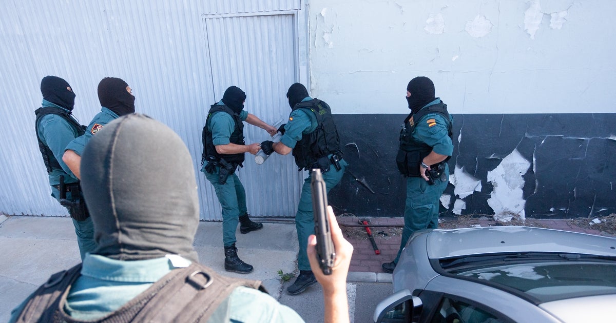 Agentes de la Guardia Civil, con pasamontañas y chalecos antibalas, realizan una intervención ante una puerta metálica, utilizando herramientas para forzarla, mientras otro agente porta una escopeta y vigila la operación. Toda la escena ocurre junto a un edificio de fachada desgastada y un coche plateado aparcado cerca, transmitiendo el ambiente de una actuación policial contra el narcotráfico.