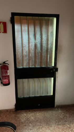 Puerta interior de cristal armado con la luna agrietada por un fuerte impacto, junto a un extintor y restos de cristal en el suelo del cuartel.