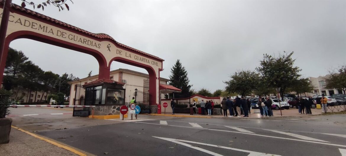 Entrada principal de la Academia de Guardias de la Guardia Civil en Baeza, con el arco de acceso rojo y crema y varios grupos de personas y vehículos congregados bajo un cielo nublado.