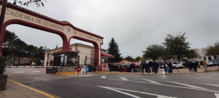 Entrada principal de la Academia de Guardias de la Guardia Civil en Baeza, con el arco de acceso rojo y crema y varios grupos de personas y vehículos congregados bajo un cielo nublado.
