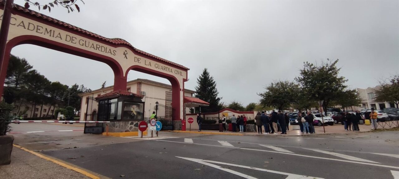 Entrada principal de la Academia de Guardias de la Guardia Civil en Baeza, con el arco de acceso rojo y crema y varios grupos de personas y vehículos congregados bajo un cielo nublado.