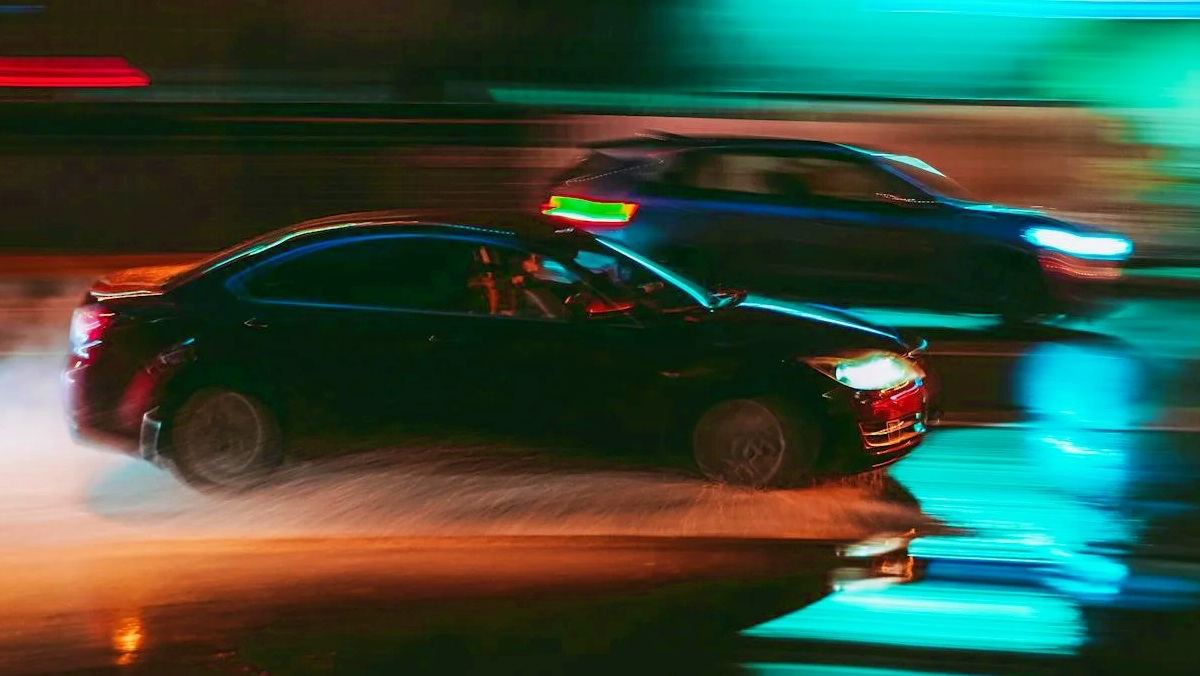 Coches circulando a gran velocidad de noche sobre calzada mojada, con luces y colores desenfocados que evocan una carrera ilegal en carretera urbana.