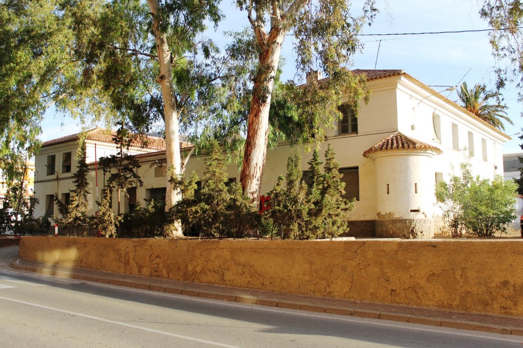 Fachada del cuartel de la Guardia Civil de Mazarrón, con un edificio blanco de dos plantas rodeado de árboles y un muro bajo junto a la calzada.
